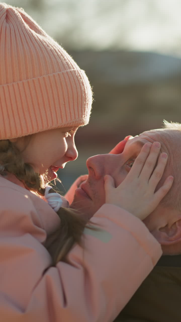 una niña alegre con una gorra rosada y una chaqueta está siendo llevada por un hombre. ella está tocando juguetón su nariz a su nariz, ambos sonriendo cálidamente y compartiendo un momento tierno y feliz al aire libre