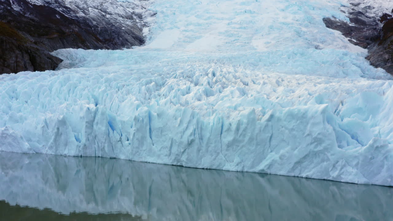 Close-up of towering blue glacier with dramatic texture and deep ridges over Patagonian sea, aerial pullback