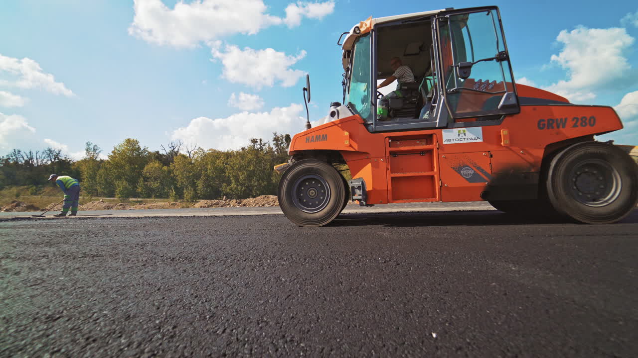 Orange roller machine pressing hot asphalt on the road outdoors. Workers and compactor machine making new asphalt. Street resurfacing.