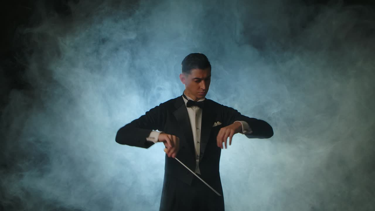 un líder de orquesta con traje usando el bastón de dirección para sincronizar a los músicos. el maestro está dirigiendo a los músicos con movimiento de palo en fondo negro con humo y luz de fondo.