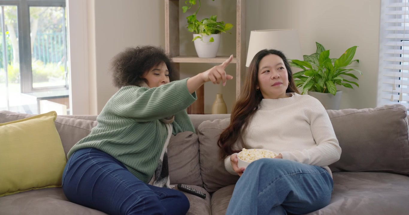 Forskellige veninder peger og reagerer på et show uden for skærmen, mens de sidder på en sofa og deler en skål popcorn.