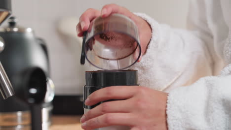 woman finishes coffee beans grinding at table closeup. lady with bath gown taps grinder cap to shake out brown powder at home. caffeine drink