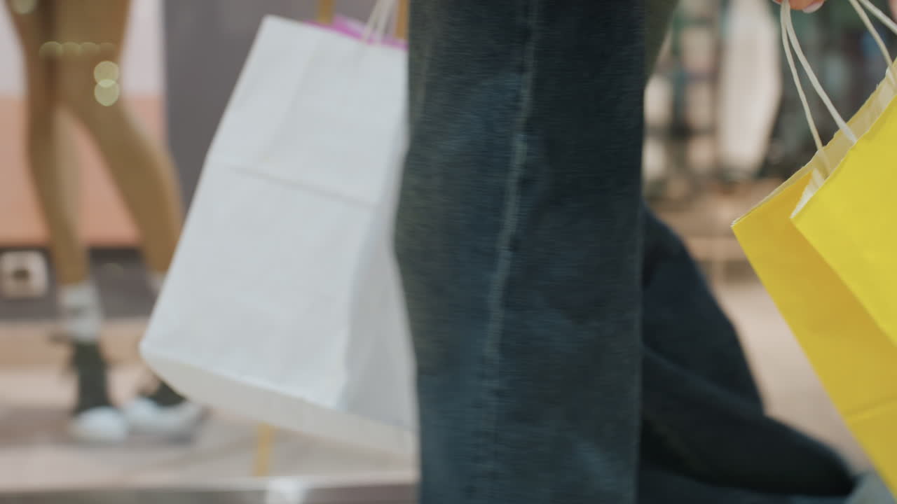 밝은 색상의 가방이 제품을 보여주는 쇼핑객 옆에서 흔들립니다. 눈에 띄는 노란색 쇼핑백이 다리와 데님 근처에서 흔들리고, 쇼핑객은 로고와 포장 세부 사항을 강조하면서 매장 환경을 돌아다닙니다.