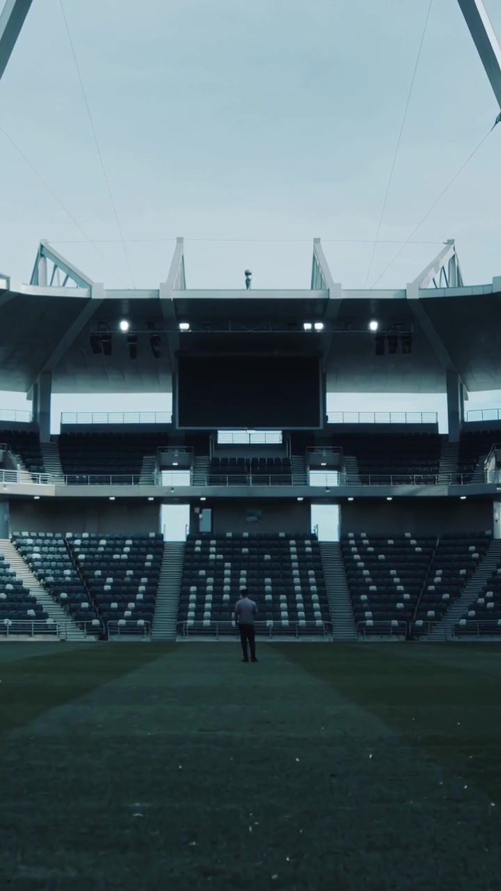 Video verticale: Uomo in piedi con cappotto scuro che si sposta mentre guarda uno schermo in uno stadio illuminato.
