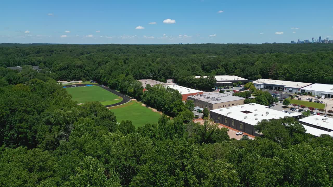 Aerial approaching shot of midtown high school with soccer field in green. Idyllic suburb area of Atlanta. Skyline with skyscraper in distance. Wide shot. Several buildings of campus. Summer sunny day