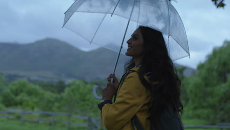 Schöne-Junge-Indische-Frau-Mit-Regenschirm-Lächelt-Glücklich-Und-Bereitet-Sich-Darauf-Vor,-Einen-Ruhigen-Regnerischen-Tag-Vor-Dem-Hintergrund-Eines-Lebendigen-Grünen-Bauernhofs-Zu-Genießen