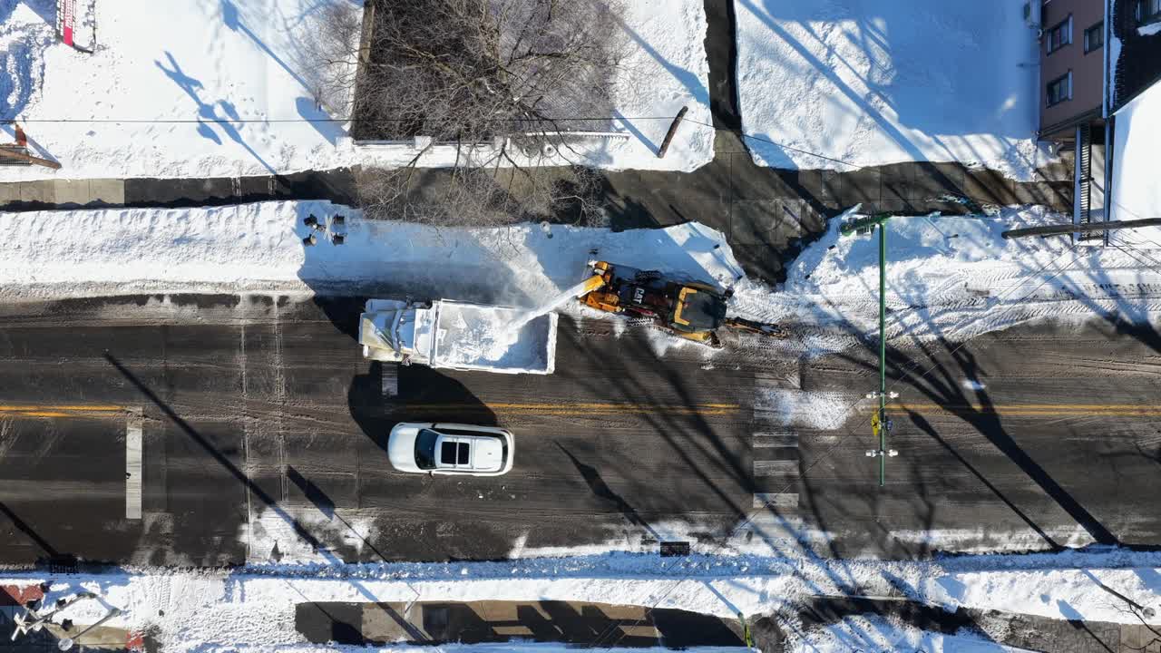 冬の米国ペンシルベニア州の静かな住宅街。除雪車が住宅街の通りを除雪し、雪が積もった歩道と家々の脇を車が通り過ぎる様子を真上から空撮。