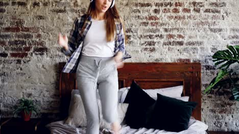 happy cheerful woman is dancing on bed at home listening to music through wireless headphones. she is wearing casual clothes, nice house interior is visible.