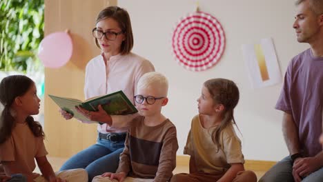 a blonde woman with a boxy hairstyle and a pink shirt reads a green book to preschool children in a special club for preparing children for school