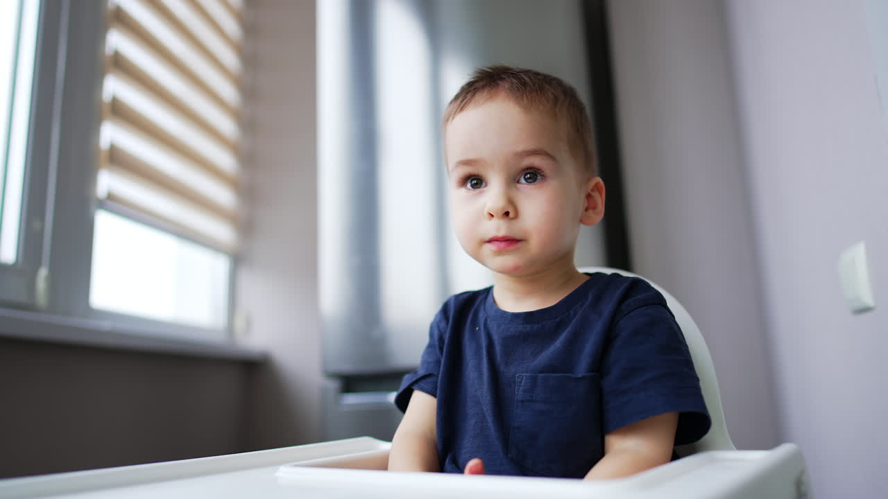 Cute Caucasian baby boy wearing dark blue t-shirt sitting at high chair. Sweet toddler waiting for his meal. Low angle view.