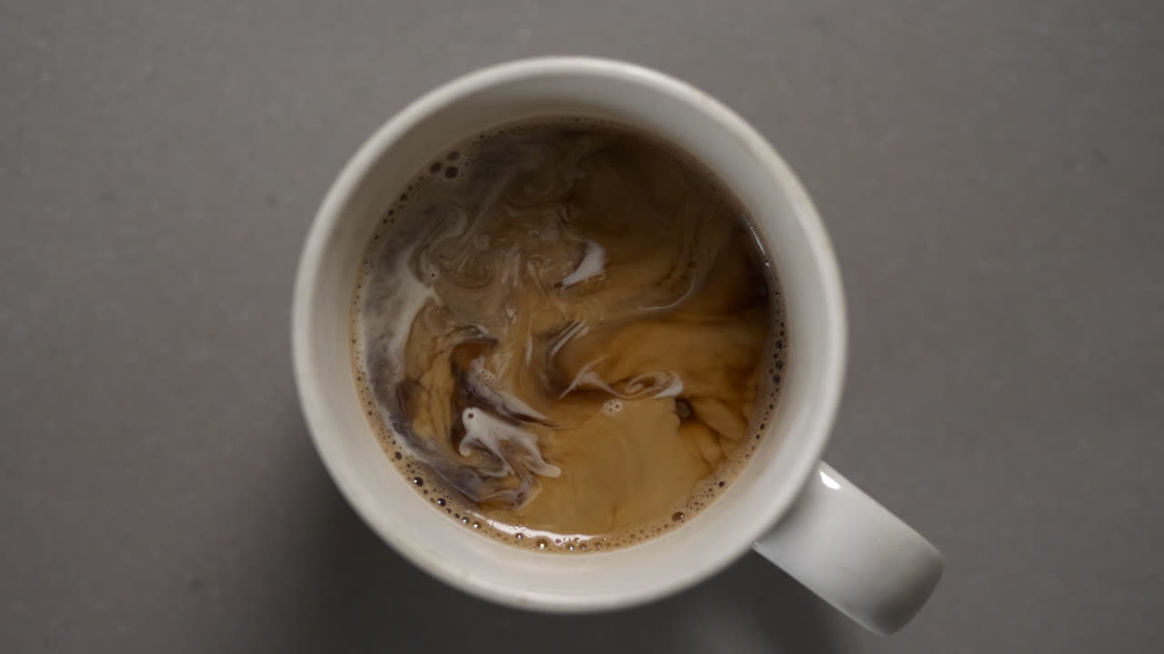 Cream or half and half dissolving into a cup of dark roasted coffee, birds eye view, slow motion, 4K