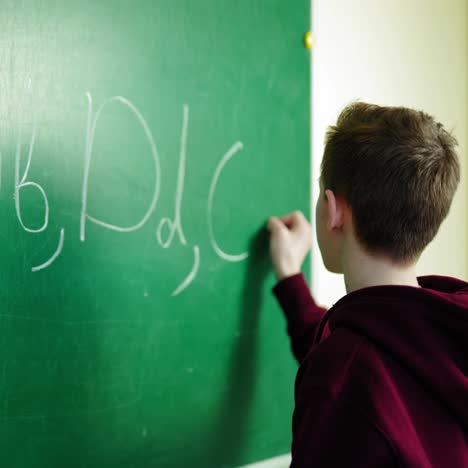 schoolboy writing on blackboard. teen boy writes alphabet on green school chalkboard. backside view of a student with a chalk in the classroom. education concept.