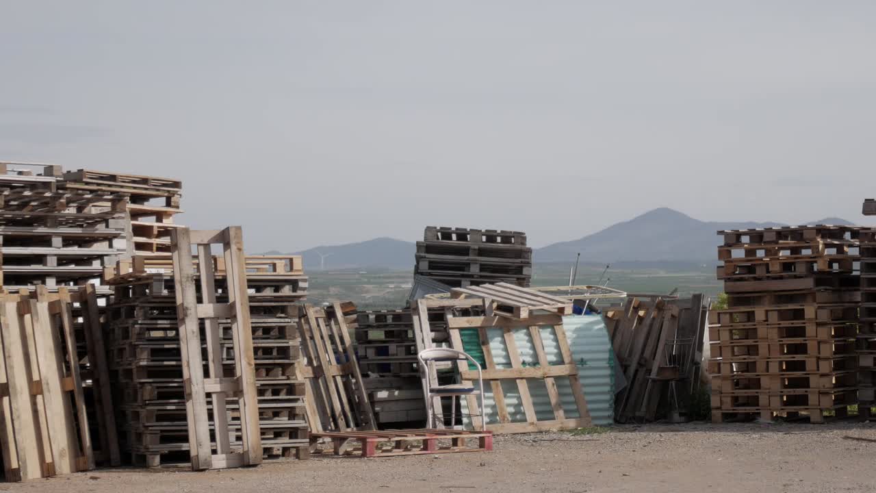 Panning shot of columns of pallets piled vertically outdoors. Different colours and sizes. Some organized others not. Horizon behind. Mountains on skyline