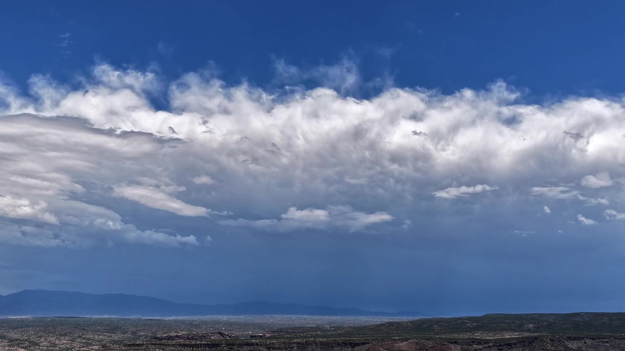 4K UHD drone-timelapse, der fanger en dramatisk, delt himmel over en barsk ørkenkløft. Et tykt bånd af lyse hvide skyer adskiller den klare øvre himmel fra et stemningsfuldt, skyggefuldt landskab.
