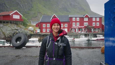 En-Cámara-Lenta,-Una-Joven-Fotógrafa-Turista-Camina-Hacia-La-Cámara-Con-Al-Fondo-Las-Casas-Rorbu-Rojas-Del-Hermoso-Pueblo-Pesquero-De-Å-En-Las-Islas-Lofoten,-Noruega