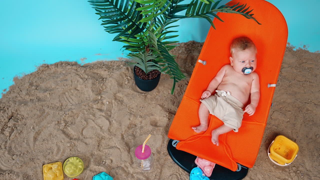 Caucasian newborn resting at the improvised beach with palm and sand. Little blond kid sitting in the orange chair. Top view.