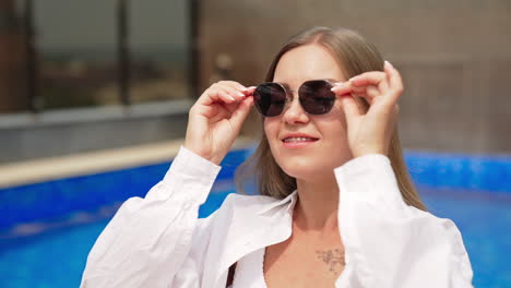 happy relaxed young lady in white shirt sitting near the pool. woman puts on her sunglasses and smiles to the camera.