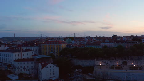 Vista-Aérea-De-Casas-Pequeñas-Tradicionales-En-El-Centro-Urbano-De-Lisboa-Con-Revelación-Del-Puente-Rojo-Ponte-25-De-Abril-Cruzando-El-Mar-En-El-Fondo