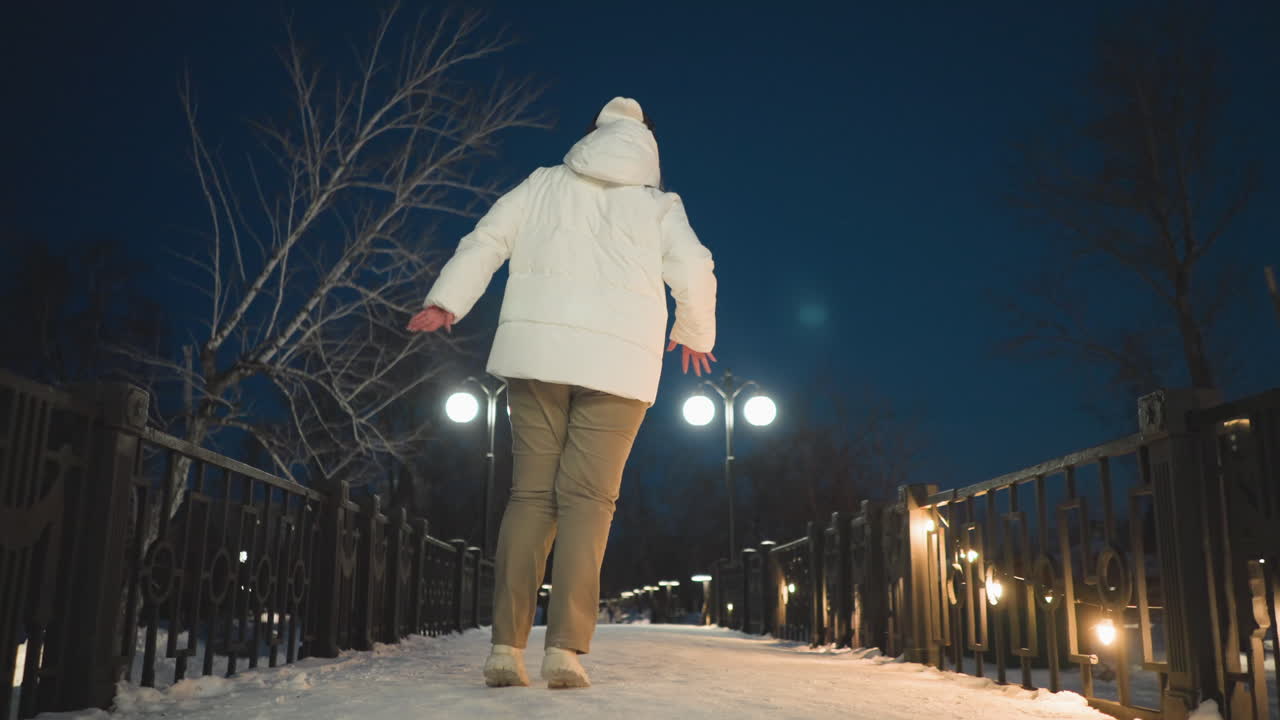Choreographer dancing gracefully in illuminated snowy park walkway wearing white puffer coat and headphones under glowing lampposts and string lights with rhythmic hand movement expressing joy