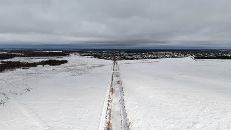 Drone-dolly-along-snow-covered-trench-road-leading-to-small-city-with-dense-clouds-on-horizon