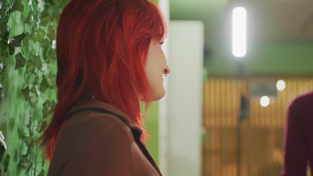 donna bianca con capelli rossi in una hall, sguardo basso e delicato, girocollo e giacca marrone, parete di piante verdi con luci calde, profilo spontaneo, che cammina tra la folla, vista posteriore casual