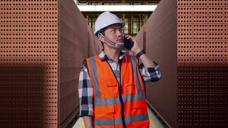 asian male engineer with safety helmet talking on smartphone while standing with red brick packed in stacks are stored