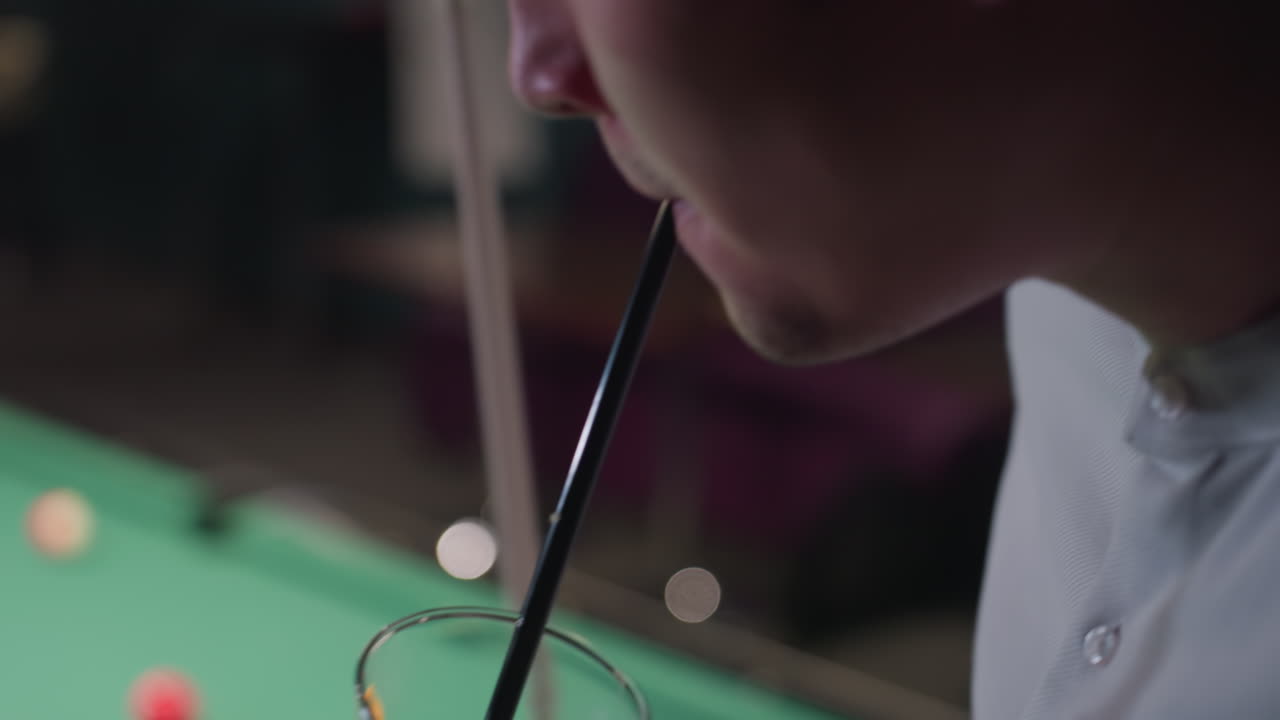 primer plano de un hombre con camisa blanca, sosteniendo un palo y un vaso de bebida de limón con paja negra. cubos de hielo y rebanada de limón flotan en el vidrio mientras lo levanta para tomar un sorbo. mesa de billar borrosa con bolas de colores