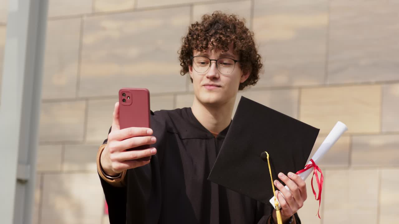 Estudante do sexo masculino estendendo um telefone vermelho para uma chamada ao vivo, segurando um barrete e um diploma perto de uma coluna de pedra