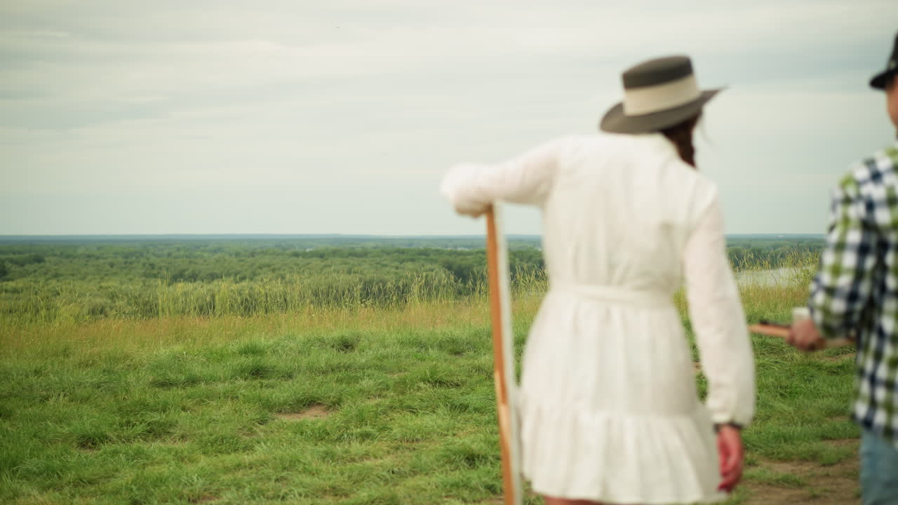 잔디에서 고 있는 한 부부의 뒷면.  옷을 입은 여자가 캔버스를 들고 있는 반면, 체크색 셔츠와 청바지를 입은 남자는 그림을 그리는 패드와 기둥을 들고 있다.