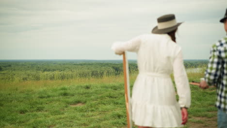 vista trasera de una pareja caminando por un campo cubierto de hierba. la mujer, con una túnica blanca, lleva un lienzo mientras que el hombre, con una camisa a cuadros y vaqueros, sostiene un bloc de dibujo y un caballete