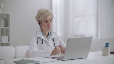 aged woman pediatrician is consulting online sitting in office of hospital and talking to web camera of laptop telemedicine session