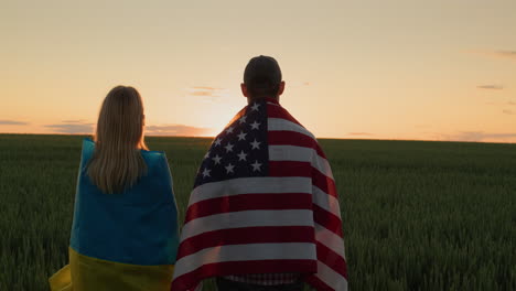 un hombre y una mujer con las banderas de ucrania y los ee.uu. se paran uno al lado del otro y miran el amanecer sobre un campo de trigo