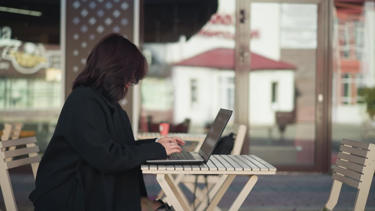 Side view of woman working on laptop outdoors with hair covering her face, seated near modern furniture in urban setting, glass building reflecting person in background, and flowers