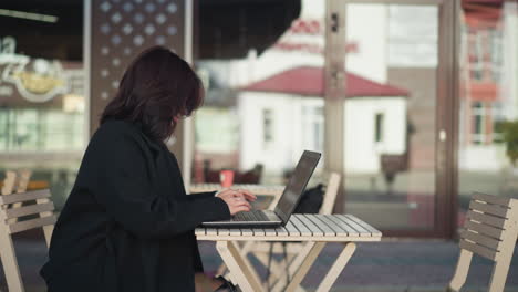 side view of woman working on laptop outdoors with hair covering her face, seated near modern furniture in urban setting, glass building reflecting person in background, and flowers