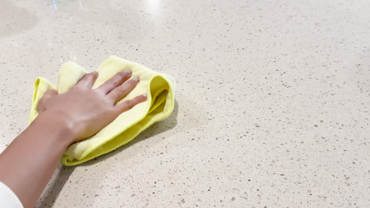 A person cleans a coffee spill on a countertop using a spray bottle and cloth under bright lighting