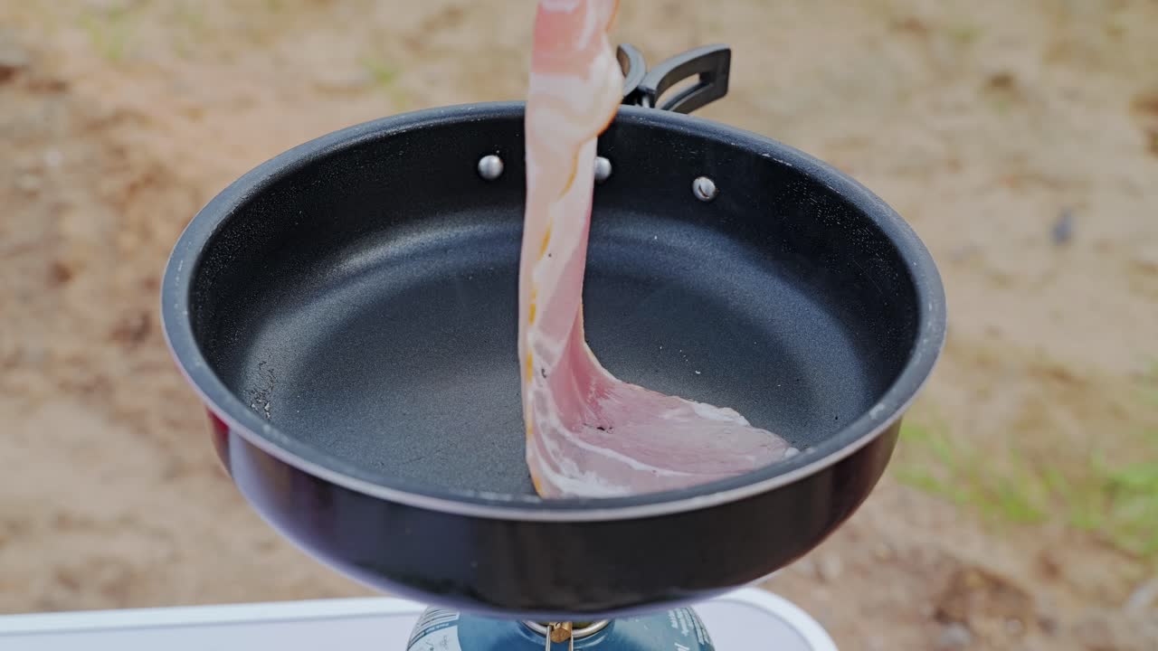 Cinematic cooking moment outdoors as bacon strip touches non-stick surface
