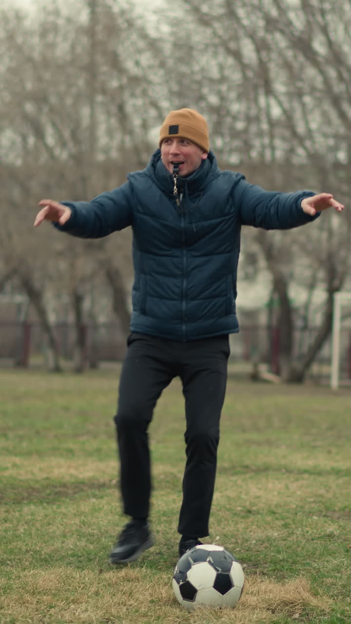 A coach sets the ball in the middle of two players facing each other as he blows the whistle, the background features a goal post, trees, and a car passing by