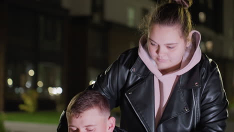 young lady in leather jacket and hoodie walks up to brother seated under night light resting gently on his shoulder while looking at him as he smiles softly with blurred urban building in background