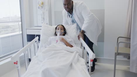 Diverse-doctor-walking-with-sick-girl-patient-wearing-oxygen-mask-in-hospital-bed-in-slow-motion