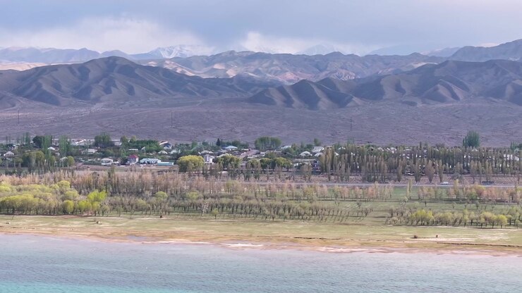 Riprese aeree del Lago Issyk Kul con catena montuosa e piccolo insediamento, paesaggio. Kirghizistan.