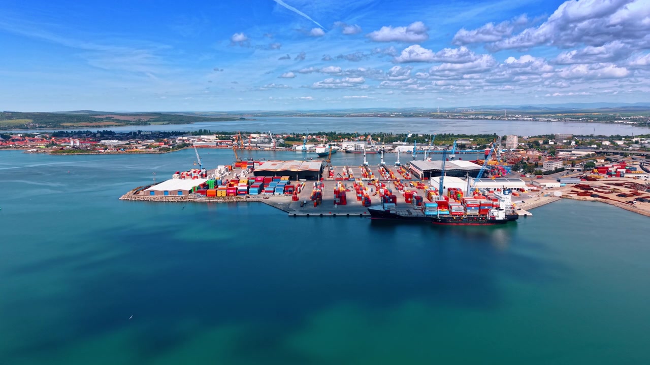 Burgas, Bulgaria, 10 aprile 2026: Terminal container e nave attraccata. Ampia angolazione di container multicolori, gru e nave con acqua calma color verde acqua e colline verdi.