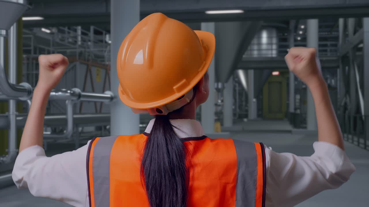 Primo piano posteriore di un ingegnere donna con casco di sicurezza che alza le mani in segno di festa mentre lavora all'interno di una distilleria con grandi serbatoi di fermentazione.