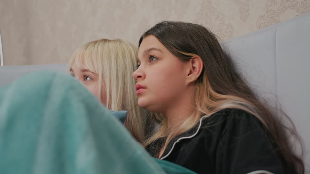two young girls sitting closely on bed wrapped in blanket while watching intense movie scene, one girl covers face with blanket, both showing expressions of fear