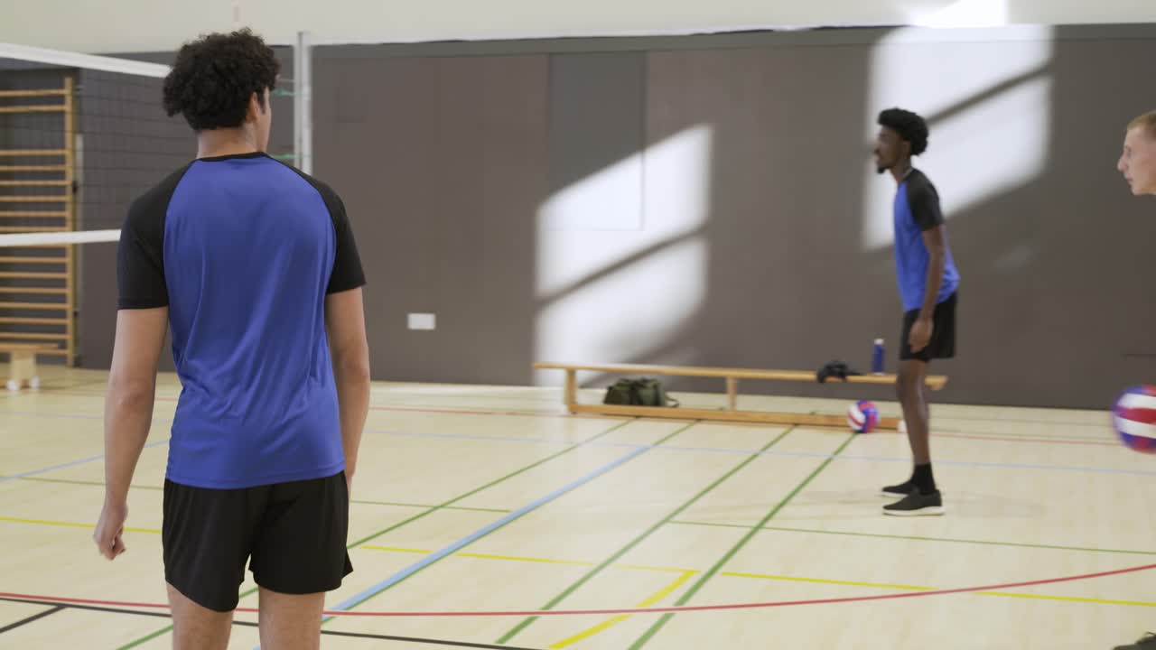 Two volleyball teams in drill; yellow serving, blue receiving, tracking ball over net in school gym