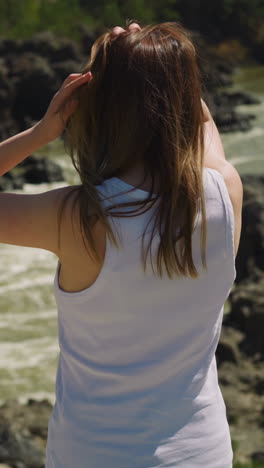 woman adjusts loose hair standing on rocky bank of mountain river on summer day backside view slow motion. tourist enjoys vacation. water flow
