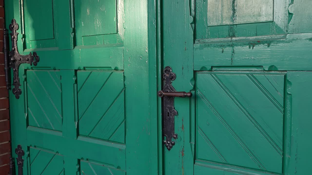 Close-up of an old green wooden door with ornate metal fittings. The camera gently moves closer to the doorknob, captured in natural light.