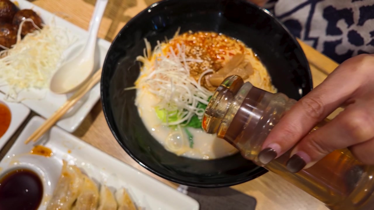 Primo piano di mani che versano olio piccante in una ciotola di cremosa zuppa di noodle giapponese