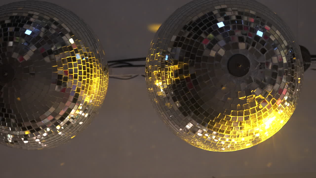 Colgando del techo, dos bolas de discoteca reflejan una luz cálida y dorada sobre un fondo gris, creando un ambiente acogedor y festivo perfecto para celebraciones y vida nocturna, plano detalle.