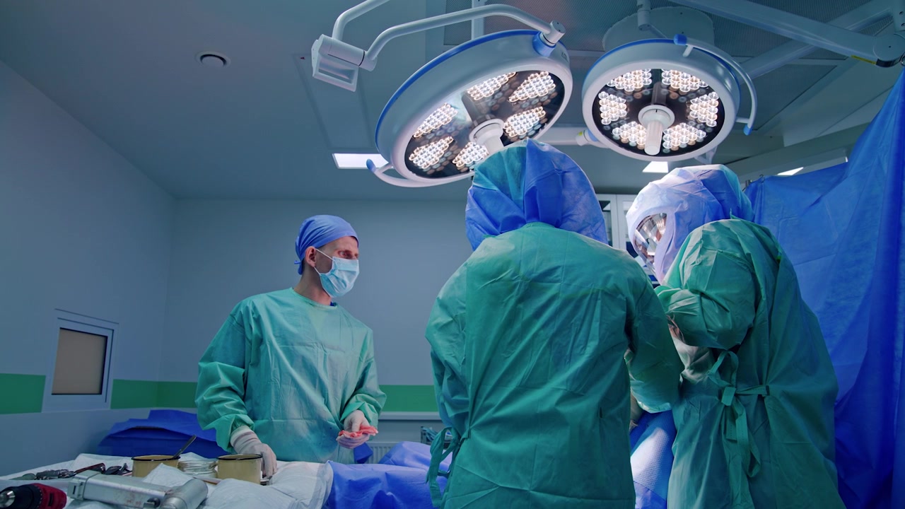 Equipe chirurgica esperta che esegue un intervento chirurgico per un trauma in una moderna sala operatoria. Un assistente maschio mascherato si trova al carrello degli strumenti