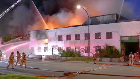 firefighters spray water to control a large fire engulfing an industrial building.